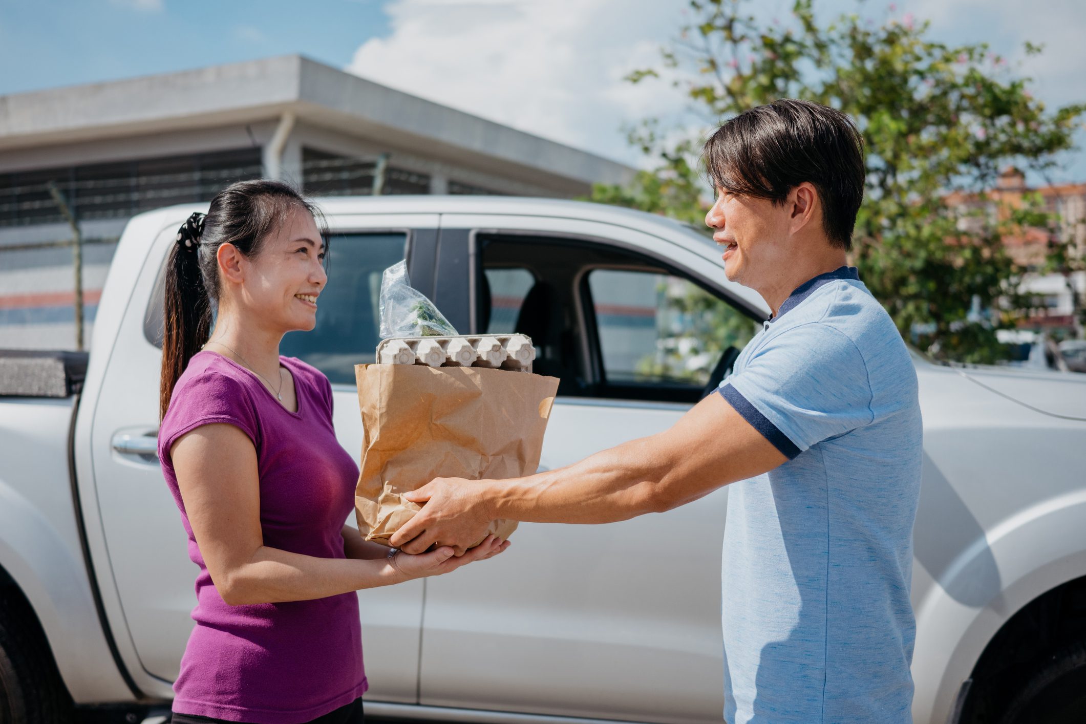 Image of an Asian delivery man delivering groceries in paper bag to woman. Mature woman receiving a bag of groceries from delivery person.