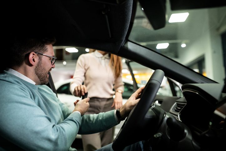 Man receives keys from saleswoman inside dealership showroom, sitting in new car and smiling with excitement and satisfaction after buying his vehicle and completing the purchase