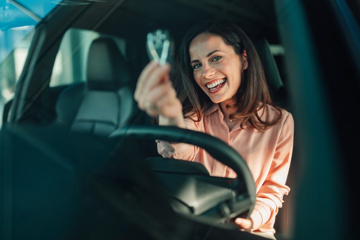 Young adult woman holding car keys, smiling happily while sitting inside a new vehicle at a dealership, celebrating a car purchase