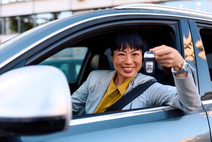 Smiling Asian mid-adult woman holds a car key outside a modern vehicle, looking at camera—celebrating ownership, rental, driving independence, commuting and business mobility