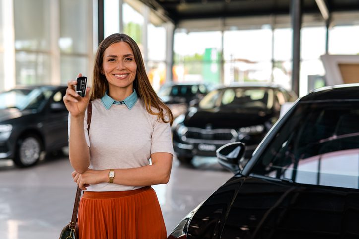 Happy woman buying a car and holding a car key in a modern car dealership showroom